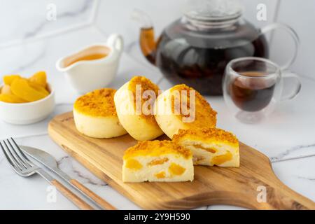 Drei weiche Käsekuchen mit goldenem Streusel, serviert auf einem Holzbrett mit Tee und Pfirsichscheiben. Stockfoto