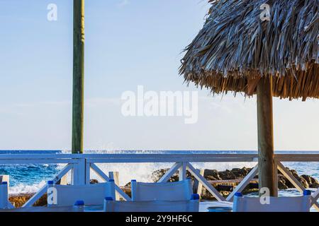 Blick auf das karibische Meer mit Wellen, die gegen Felsen plätschern, von unter dem Strohdach der Cabana an sonnigem Tag gesehen. Curacao. Stockfoto