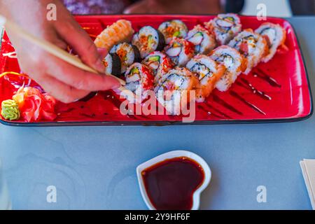 Nehmen Sie Sushi von der farbenfrohen Platte mit verschiedenen Sushi-Brötchen und herzförmiger Sojasauce von Hand mit Essstäbchen auf. Stockfoto