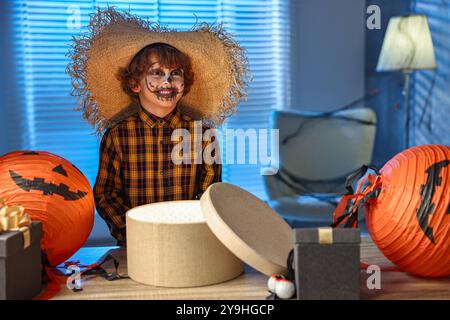 Süßer Junge, der wie Vogelscheuche gekleidet ist, mit festlichem Dekor und Geschenkboxen drinnen in der Nacht. Halloween-Feier Stockfoto