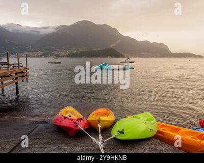 Farbenfrohe Kajaks liegen am ruhigen Ufer des Comer Sees nahe Lezzeno, mit Booten und Bergen im Hintergrund bei Sonnenuntergang. Stockfoto