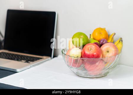 Eine klare Glasschale gefüllt mit verschiedenen Früchten, darunter Äpfel, Bananen, eine Limette, eine Birne und eine Pflaume, auf einer weißen Tischdecke neben einem Laptop platziert. Stockfoto