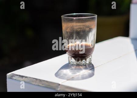 Ein Glas mit dunkler Flüssigkeit, möglicherweise Kaffee, steht auf einer weißen Oberfläche. Das Glas ist teilweise gefüllt und zeigt Reste des Getränks. Der Hintergrund Stockfoto