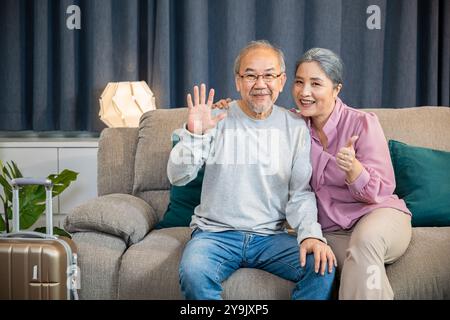 Glückliches asiatisches Seniorenpaar, das jemanden begrüßt, während er auf einer bequemen Couch sitzt, alter Mann und Frau Stockfoto