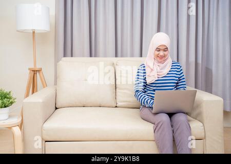 Asiatische muslimische Frau im Hijab sitzend mit dem Laptop zur Arbeit, zu Hause auf dem Sofa zu arbeiten und fernzusehen. Stockfoto