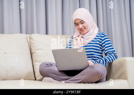 Asiatische muslimische Frau im Hijab sitzend mit dem Laptop zur Arbeit, zu Hause auf dem Sofa zu arbeiten und fernzusehen. Stockfoto