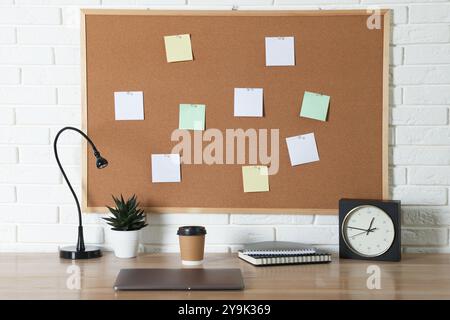 Korkbrett mit leeren Notizen, Laptop, Lampe, Wecker und Notizbüchern auf Holztisch in der Nähe der weißen Ziegelwand Stockfoto