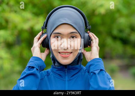 Asiatische süße muslimische Frau in Sportbekleidung und blauem Hijab, die im Park draußen Musik auf ihrem Telefon hört Stockfoto