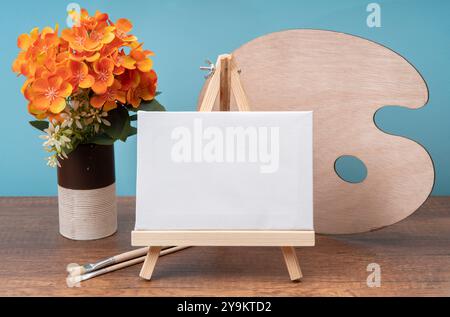Vorderansicht der kleinen Holzstaffelei mit leerer Leinwand auf Holztisch, dekoriert mit Glas mit Blumen, Pinseln und Palette vor blauem Hintergrund Stockfoto