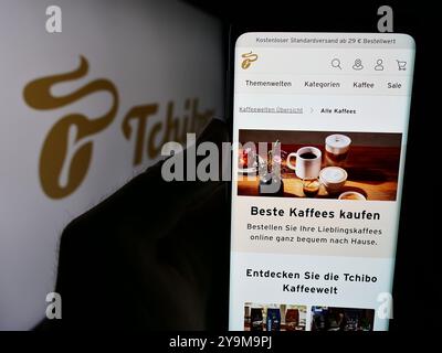 In dieser Abbildung hält eine Person ein Smartphone mit der Webseite des deutschen Kaffeehändler Tchibo GmbH vor dem Firmenlogo. Stockfoto