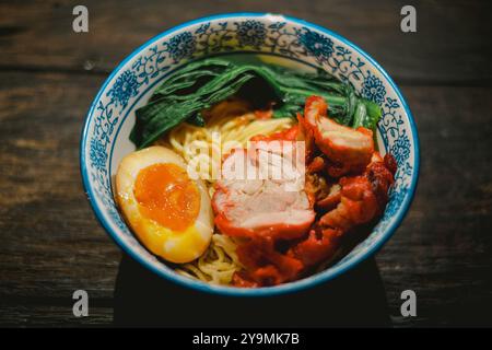 Chinesische gebratene Nudeln in einer Schüssel mit Char Siu, hart gekochtem Ei und Gemüse auf Holztisch. Kantonesische Küche im kopitiam-Stil. Draufsichtswinkel Stockfoto