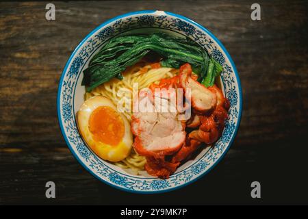 Chinesische gebratene Nudeln in einer Schüssel mit Char Siu, hart gekochtem Ei und Gemüse auf Holztisch. Kantonesische Küche im kopitiam-Stil. Flacher Lagewinkel Stockfoto