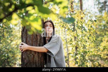 Eine Frau lehnt sich an einen Baum, blickt mit einem ruhigen Ausdruck nach oben, umgeben von leuchtend grünen Blättern in einem sonnendurchfluteten Wald. Stockfoto