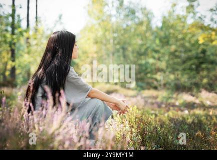 Eine junge Frau mit langen Haaren sitzt auf einem Feld, in Gedanken verloren, umgeben von blühenden Wildblumen unter dem warmen Sonnenlicht in einer ruhigen Waldlandschaft Stockfoto