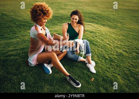 Zwei College-Freunde feiern mit Champagner, sitzen auf Gras und genießen einen sonnigen Tag im Freien mit Lachen und Freude. Stockfoto