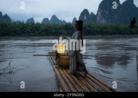 Szene von Hanfu Girl auf Bambusfloß, das mit geöffneten Armen zur blauen Stunde mit chinesischer Laterne posiert Stockfoto