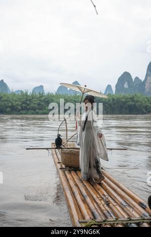 Eine Frau in Hanfu-Kleidung hält einen Schirm, während sie auf einem Bambusfloß in Xingping, China, steht Stockfoto