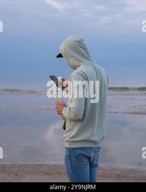 Junger Mann mit Kapuzenpullover, der am Strand steht. Er hat Handy in einer Hand und Trinkflasche in der anderen. Er guckt auf das Handy. Rückansicht. Stockfoto