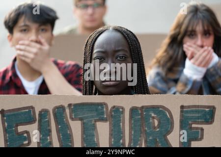 Gruppe verschiedener junger Menschen bei einer Demonstration zum Klimawandel, mit einer ernsten Afrikanerin im Fokus, die ein Zeichen für DIE ZUKUNFT hält. Andere decken ihre mou ab Stockfoto