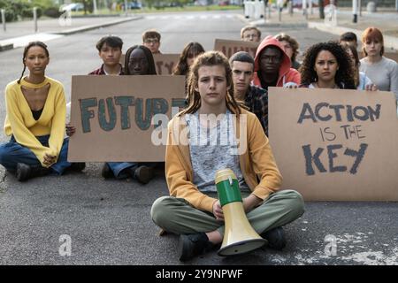 Eine Gruppe verschiedener junger Menschen, die an einem Sit-in-Protest teilnehmen und Schilder mit der Aufschrift "Zukunft" und "Aktion ist der Schlüssel" halten. Das Bild fängt Jugendaktivismus ein Stockfoto