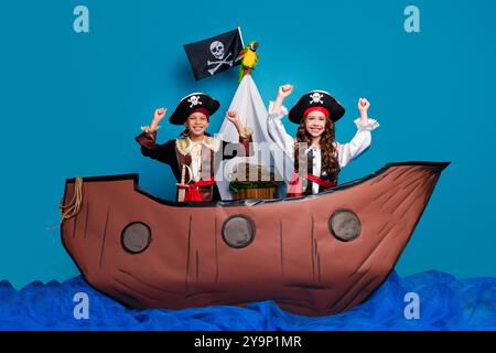 Foto in voller Länge von zwei kleinen Kindern heben Fäuste, die gekleidete Piratenkostüme gewinnen Geburtstagsparty spielen Schiff isoliert auf blauem Hintergrund Stockfoto