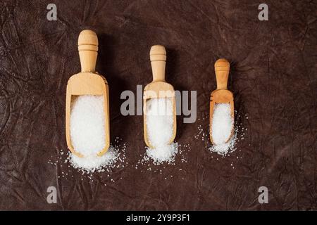 Drei hölzerne Schaufeln in verschiedenen Größen voller weißem Kristallzucker auf braunem Lederhintergrund, Bonbons Portion Konzept Stockfoto