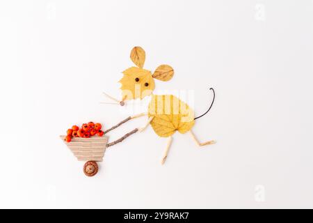 Maus aus gelben Herbstblättern, die eine Schubkarre mit Beeren schiebt, minimalistische flache Komposition mit Kopierraum. Herbst- und Naturhandwerk Konz Stockfoto