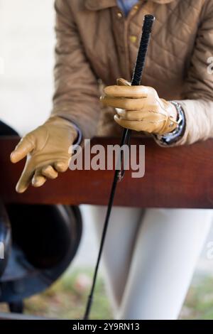 Eine Frau in beigefarbenen Handschuhen und Jockeyhosen mit einer Peitsche in den Händen steht in einer stabilen Nahaufnahme neben einem Sattel Stockfoto