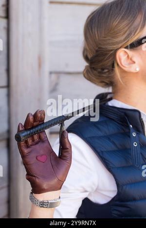 Eine Frau in braunen Handschuhen und einer Peitsche in den Händen steht in einem Stall, Nahaufnahme Stockfoto