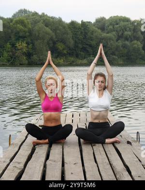 Zwei Mädchen im Teenageralter machen Yoga-Übungen zur Konzentration und Entspannung auf einem hölzernen Pier am Fluss. Das Konzept der körperlichen und psychischen Gesundheit des Teenagers Stockfoto