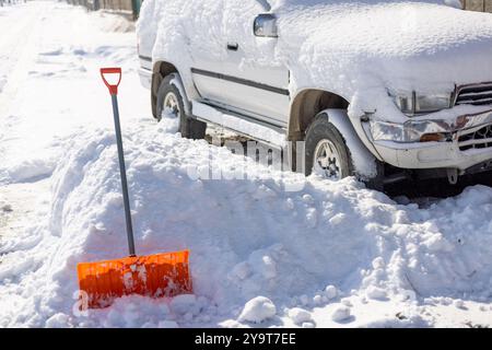 Die orangefarbene Schneeschaufel ragt aus der Schneewolke vor dem schneebedeckten weißen SUV-Auto hervor Stockfoto