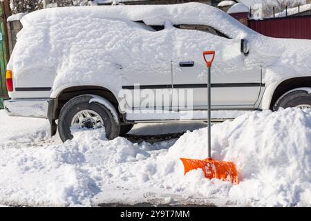 Die orangefarbene Schneeschaufel ragt aus der Schneewolke vor dem schneebedeckten weißen SUV-Auto hervor Stockfoto