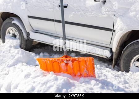 Die orangefarbene Schneeschaufel ragt aus der Schneewolke vor dem schneebedeckten weißen SUV-Auto hervor Stockfoto