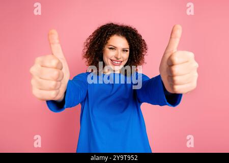 Die lächelnde junge Frau mit lockigem Haar zeigt einen doppelten Daumen nach oben vor einem fröhlichen rosa Hintergrund Stockfoto