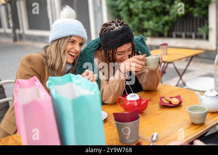 Zwei verschiedene Frauen genießen Tee und lachen im Café im Freien nach dem Einkaufen. Blonde Frau in Mütze und afroamerikanischer Freund mit Zöpfen teilen sich freudig Stockfoto