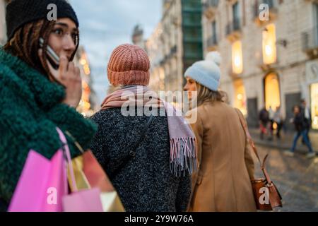 Drei junge Frauen, die abends in der beleuchteten Stadtstraße einkaufen. Freunde verschiedener Ethnien, die Taschen tragen, gehen zusammen in wi Stockfoto
