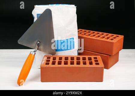 Zementpulver in Beutel, Kelle und roten Ziegeln auf weißem Holztisch. Baumaterialien und Bauwerkzeuge Stockfoto