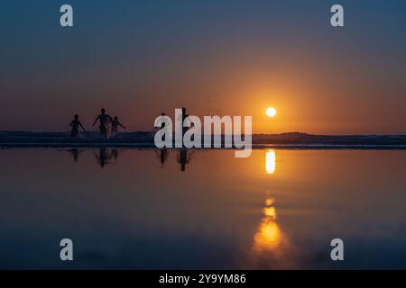 Silhouetten von Menschen, die in die Wellen laufen, während die Sonne über dem Meer untergeht, ihre Reflexionen auf dem nassen Sand glitzern. Panoramafoto Stockfoto