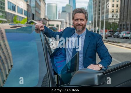Foto eines reiferen Geschäftsmannes in der Nähe des Autos. Glücklicher Geschäftsmann posiert auf seinem neuen Auto. Erfolgsgeschäft. Auto für Geschäftsleute. Gutaussehender Mann im Anzug sitzt stehend Stockfoto