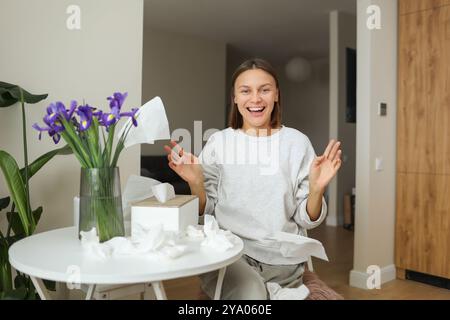 Allergiefrei. Eine fröhliche junge Frau wirft zu Hause Servietten hoch. Glückliches Leben ohne laufende Nase, Juckreiz oder Husten Symptome. Allergenstrauß von Irisblumen in der Vase, benutzte Taschentücher auf dem Tisch. Stockfoto