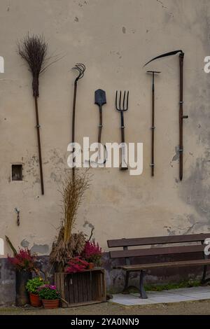 Alte Landwirtschaftsgeräte hängen an einer rustikalen Wand mit Topfpflanzen und einer Bank. Das Konzept der traditionellen Landwirtschaft, des ländlichen Lebensstils und des Handwerks Stockfoto