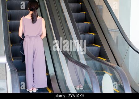 Eine Frau in einem hellvioletten Jumpsuit steht auf einer Rolltreppe, nach oben gerichtet. Die Rolltreppe hat ein modernes Design mit sichtbaren gelben Sicherheitsmarkierungen. Die Stockfoto