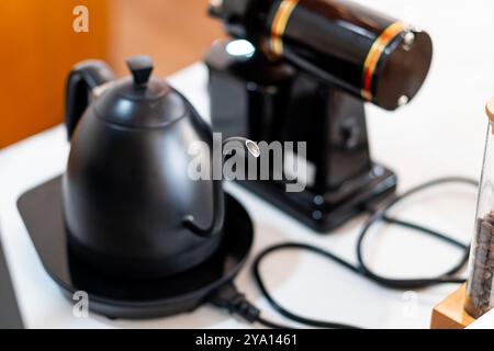 Ein eleganter schwarzer Wasserkocher mit langem Auslauf befindet sich auf einem schwarzen Sockel neben einer Kaffeemühle. Der Wasserkocher hat ein modernes Design, und das Schleifgerät verfügt über ein Schienbein Stockfoto