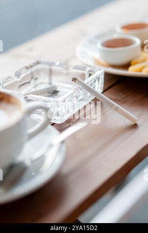 Eine gemütliche Cafeszene mit einer Tasse Kaffee, einem Aschenbecher mit einer Zigarette und einem Teller mit Snacks. Die Umgebung ist warm und einladend, perfekt zum Entspannen Stockfoto
