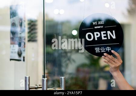 Eine Person mit einem offenen Schild vor einer Glastür, das anzeigt, dass das Unternehmen geöffnet ist. Das Schild enthält sowohl englischen als auch chinesischen Text mit einem Stockfoto