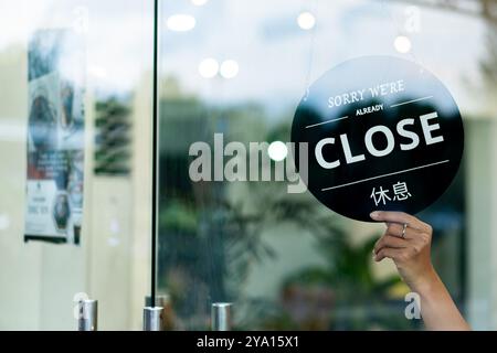 Eine Hand, die ein rundes schwarzes Schild hält, auf dem steht "SORRY WE're are CLOSE" auf Englisch und Chinesisch. Das Schild wird an einer Glastür angezeigt, was darauf hinweist Stockfoto