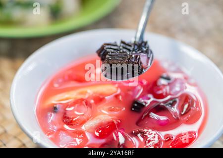 Nahaufnahme einer Schüssel mit farbenfrohen Desserts, mit Gelee-Würfeln und verschiedenen Früchten in einem süßen Sirup. Ein Löffel hebt ein Stück schwarzer Gelee Stockfoto