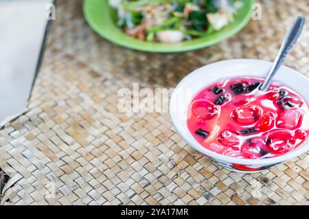Eine Schüssel mit farbenfrohen Desserts mit roten Gelee-ähnlichen Kugeln und schwarzen Streifen, serviert mit einem Löffel. Der Hintergrund zeigt eine grüne Platte mit Salat, alles Pl Stockfoto