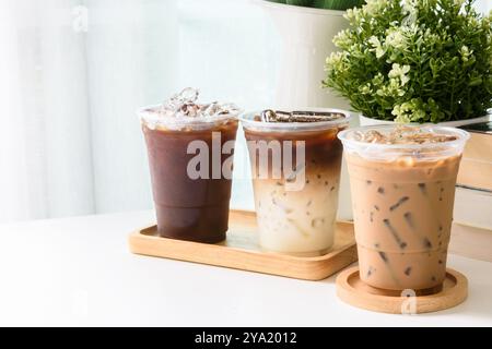 Ice americano, Ice Espresso, Ice Latte Coffee und ein Holztablett auf einem weißen Tisch im Innenbereich mit weißen Vorhängen und Blumen. Stockfoto