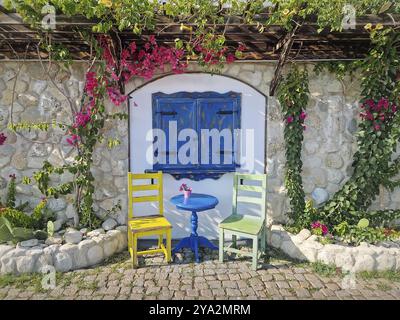 Ästhetisches Café im Hintergrund. Malerische Umgebung im Freien mit einem blauen Holzfenster, zwei farbenfrohen Stühlen und einem kleinen runden Tisch, Steinmauer, verziert Stockfoto